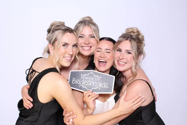 Bride and three bridesmaids smiling around a Here for the Bridesmaids sign.