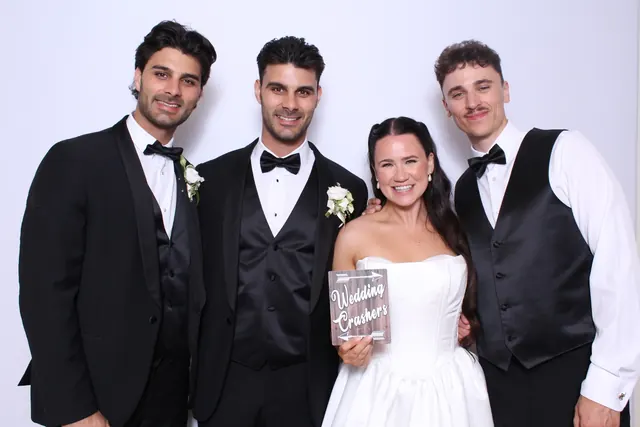 Bride posing with three formally dressed wedding guests against a white photo booth backdrop.