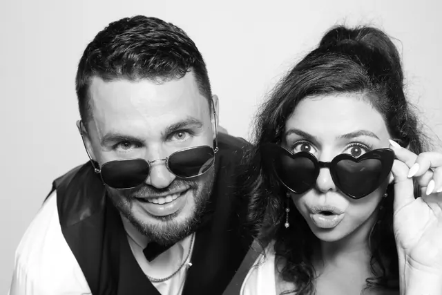 Two guests peeking over novelty sunglasses in a black-and-white Glam photo booth portrait.