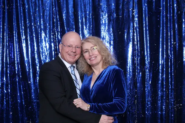 Formally dressed couple smiling in front of a blue sequin photo booth backdrop.