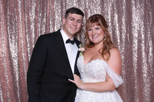 Newlyweds smiling together in front of a rose-gold sequin photo booth backdrop.