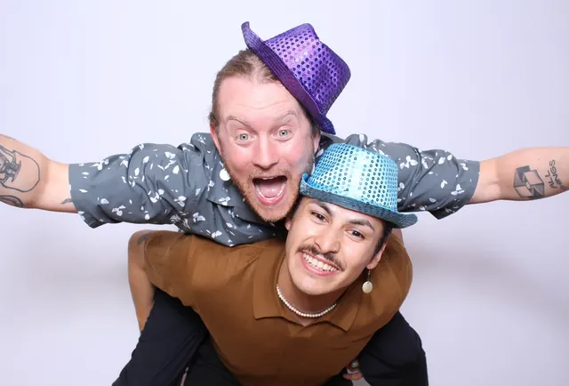 Two guests wearing colourful hats in a playful piggyback pose against a white backdrop.
